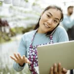Young Attractive Asian Woman Working at the Plants Nursery Using Smartphone and Laptop
